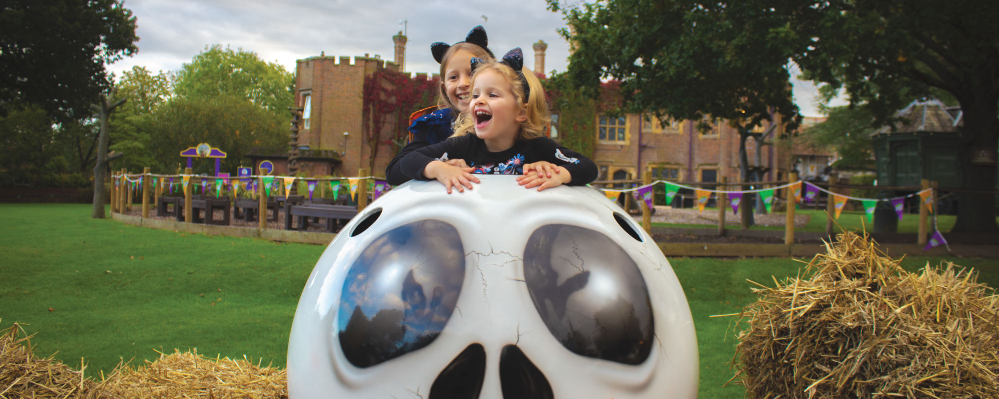 2 young girls climbing on a large skeleton head dressed up as cats for halloween.